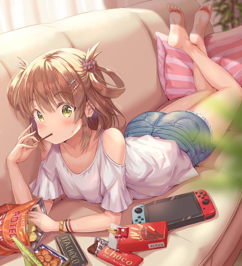 1girl, bare_shoulders, barefoot, blouse, blush, bracelet, breasts, brown_hair, candy, chips_(food), chocolate, chocolate_bar, collarbone, couch, crossed_legs, denim, denim_shorts, eating, food, green_eyes, hair_ornament, hair_rings, hairpin, handheld_game_console, highres, hirokazu_(analysis-depth), jewelry, looking_at_viewer, lying, nintendo_switch, no_shoes, on_stomach, pillow, pocky, potato_chips, princess_connect!, rino_(princess_connect!), rino_(real)_(princess_connect!), shirt, shorts, small_breasts, soles, solo, the_pose, two_side_up, white_shirt, wristband