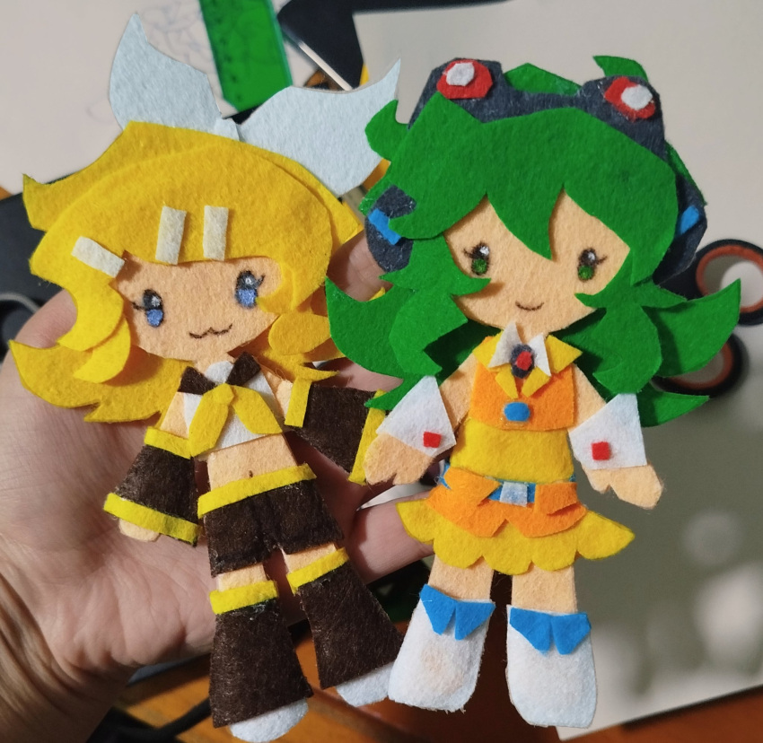 2girls, :3, art_tools_in_frame, black_shorts, blonde_hair, blue_eyes, bow, closed_mouth, commentary, critinikaqwq, deformed, detached_sleeves, dress, english_commentary, felt_(medium), full_body, goggles, goggles_on_head, green_eyes, green_hair, gumi, hair_bow, hair_ornament, hairclip, headphones, highres, kagamine_rin, leg_warmers, looking_at_viewer, medium_hair, midriff, multiple_girls, neckerchief, orange_dress, photo_(medium), shirt, shorts, smile, unconventional_media, vocaloid, white_bow, white_shirt, yellow_neckerchief