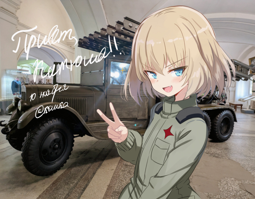 1girl, absurdres, blonde_hair, blue_eyes, bm-13_katyusha, bob_cut, commentary, cyrillic, fang, girls_und_panzer, green_jumpsuit, highres, indoors, jumpsuit, katyusha_(girls_und_panzer), koshka_pic, long_sleeves, looking_at_viewer, military_truck, military_vehicle, monkey_girl, motor_vehicle, multiple_launch_rocket_system, name_connection, open_mouth, pravda_military_uniform, russian_text, short_hair, smile, solo, standing, translation_request, truck, v